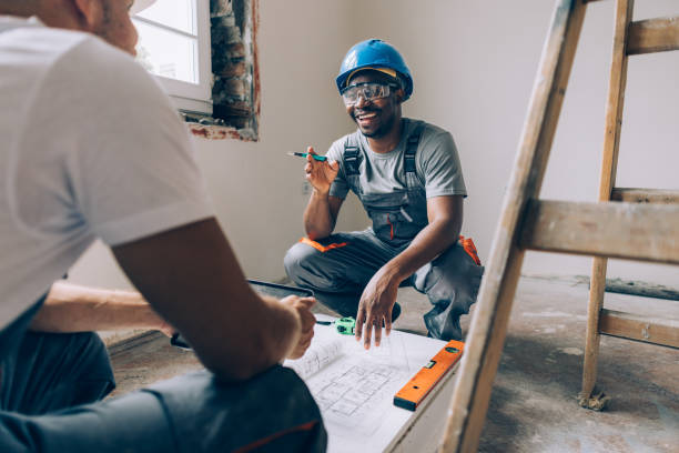 Двое строителей обсуждают чертежи, один улыбается и сидит на полу в защитном шлеме в ремонтируемой комнате.