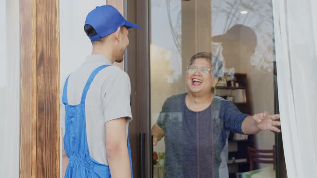 Сотрудник службы доставки в синей кепке и комбинезоне вручает посылку жизнерадостной пожилой женщине у ее входной двери.