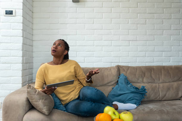 Женщина выглядит озадаченной, держа планшет и сидя на диване с вазой с фруктами на столе в гостиной с белым